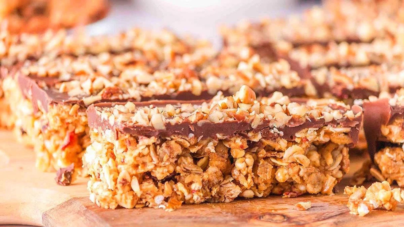 Granola bars topped with chocolate and chopped nuts, arranged on a wooden surface.