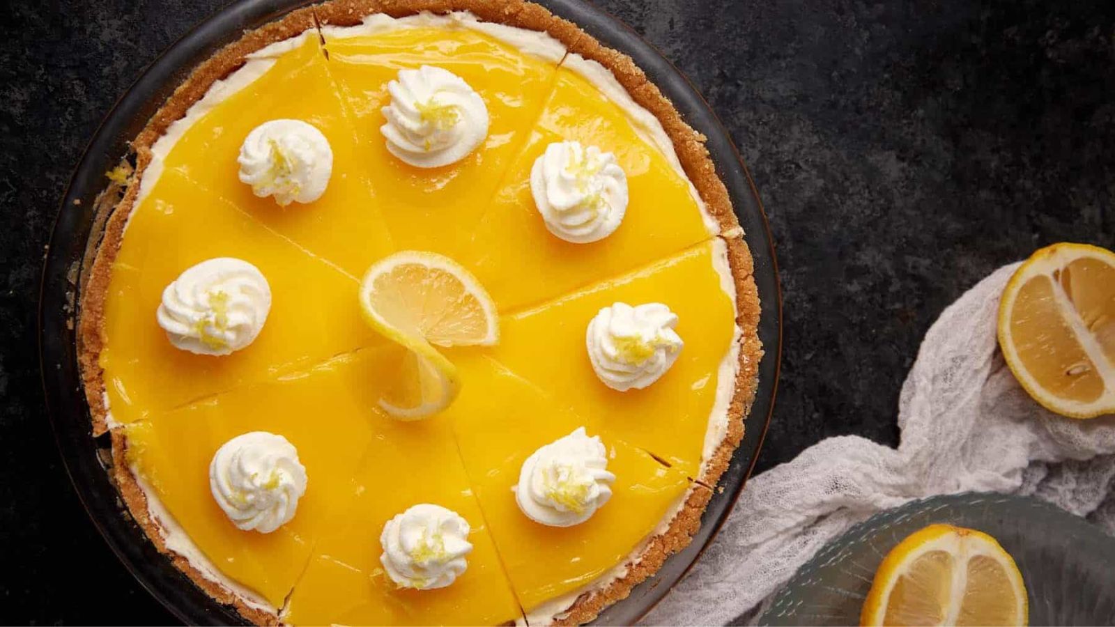 A lemon pie topped with whipped cream dollops and a lemon slice, sliced and ready to serve.