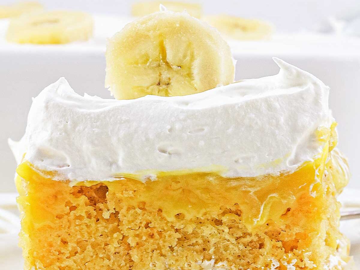 A slice of banana cream poke cake topped with whipped cream and a banana slice on a white plate.