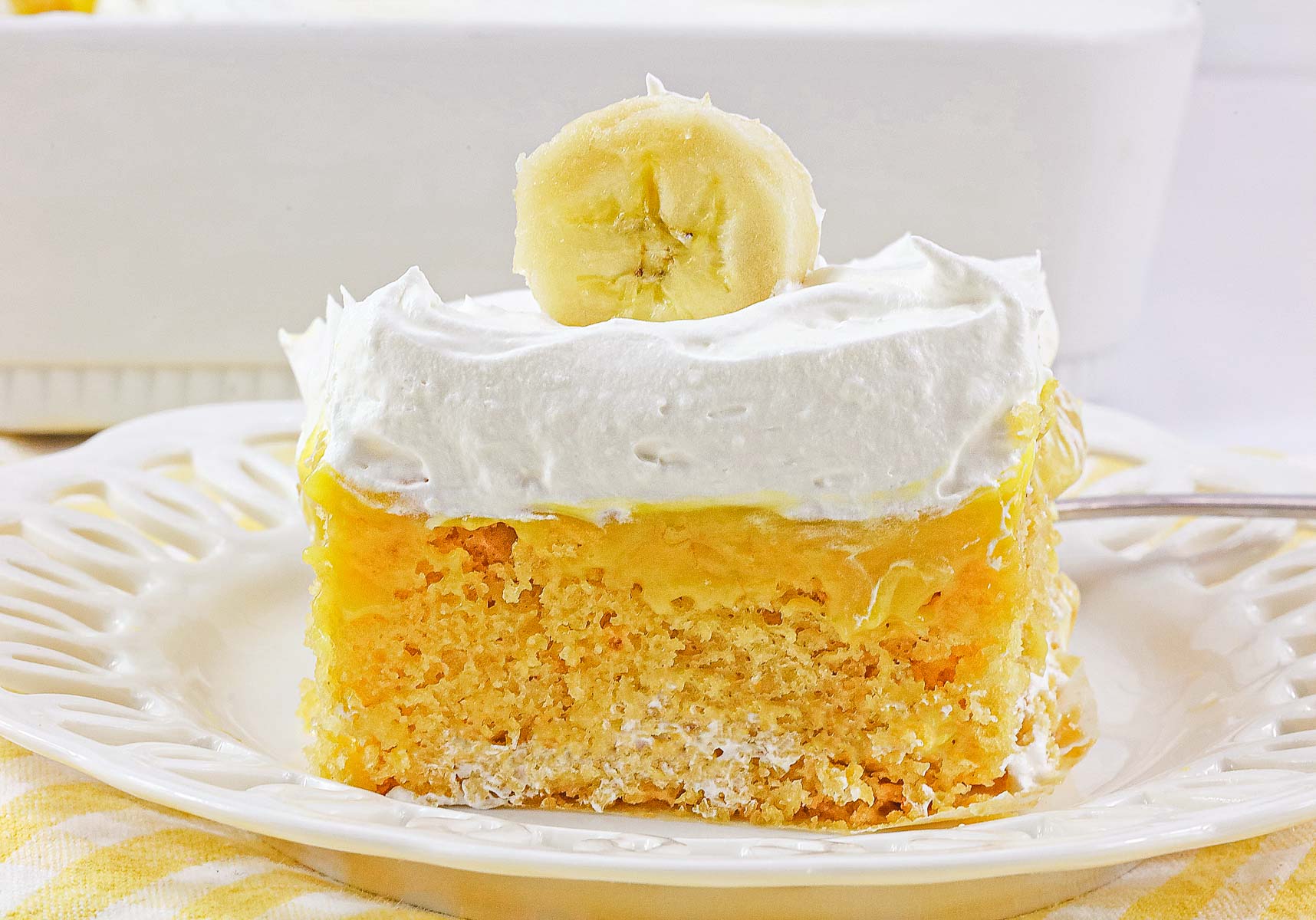 A slice of banana cream poke cake with whipped topping and a banana slice on a white plate—this easy no egg dessert is pure comfort.