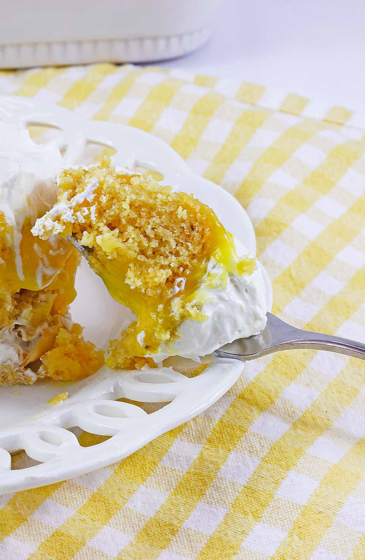 A fork holds a bite of lemon poke cake with whipped cream on a white plate, set atop a yellow checkered cloth.