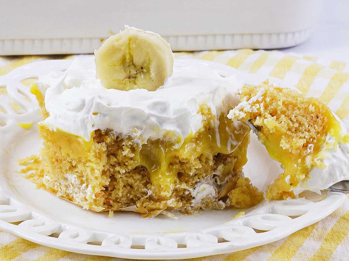 A slice of no egg banana pudding poke cake with whipped topping and a banana slice sits on a white plate, with a forkful taken out.
