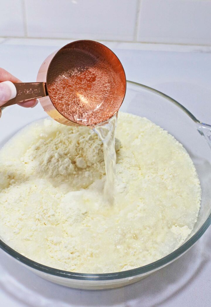 A hand pours water from a copper measuring cup into a bowl of flour, preparing the base for a no egg banana cream poke cake.