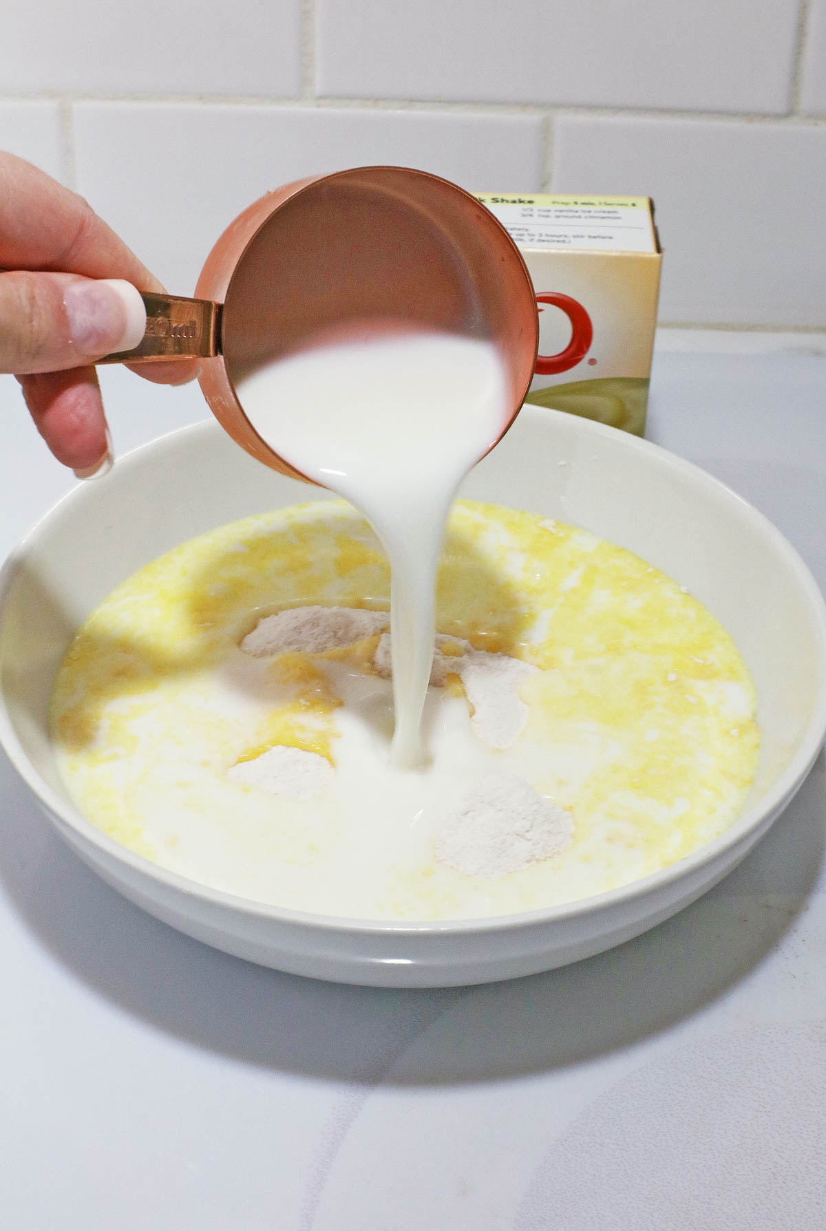 A hand pours milk from a measuring cup into a bowl with flour for a No Egg banana cream poke cake recipe on a kitchen counter.