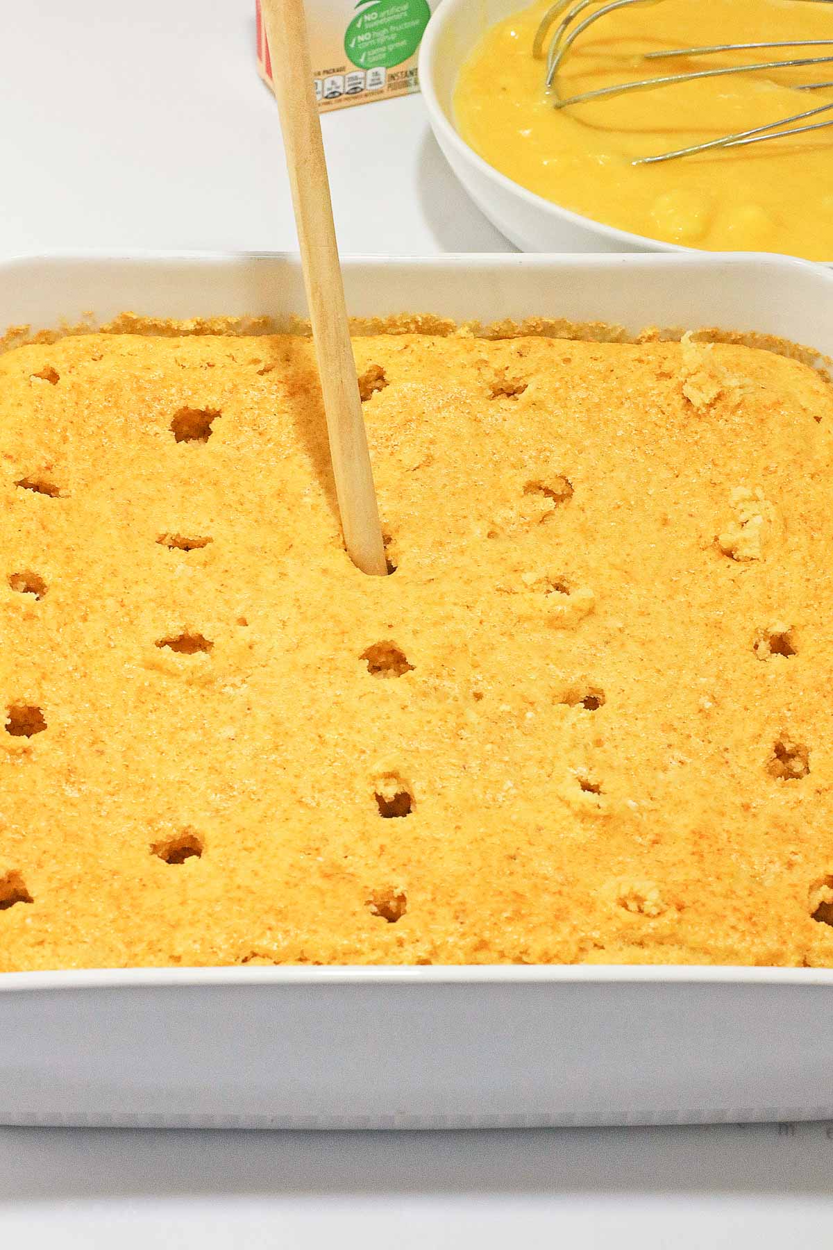 A baked Banana Cream poke cake in a white dish with holes poked in it, next to a bowl of yellow pudding and a whisk—perfect for a no egg dessert.