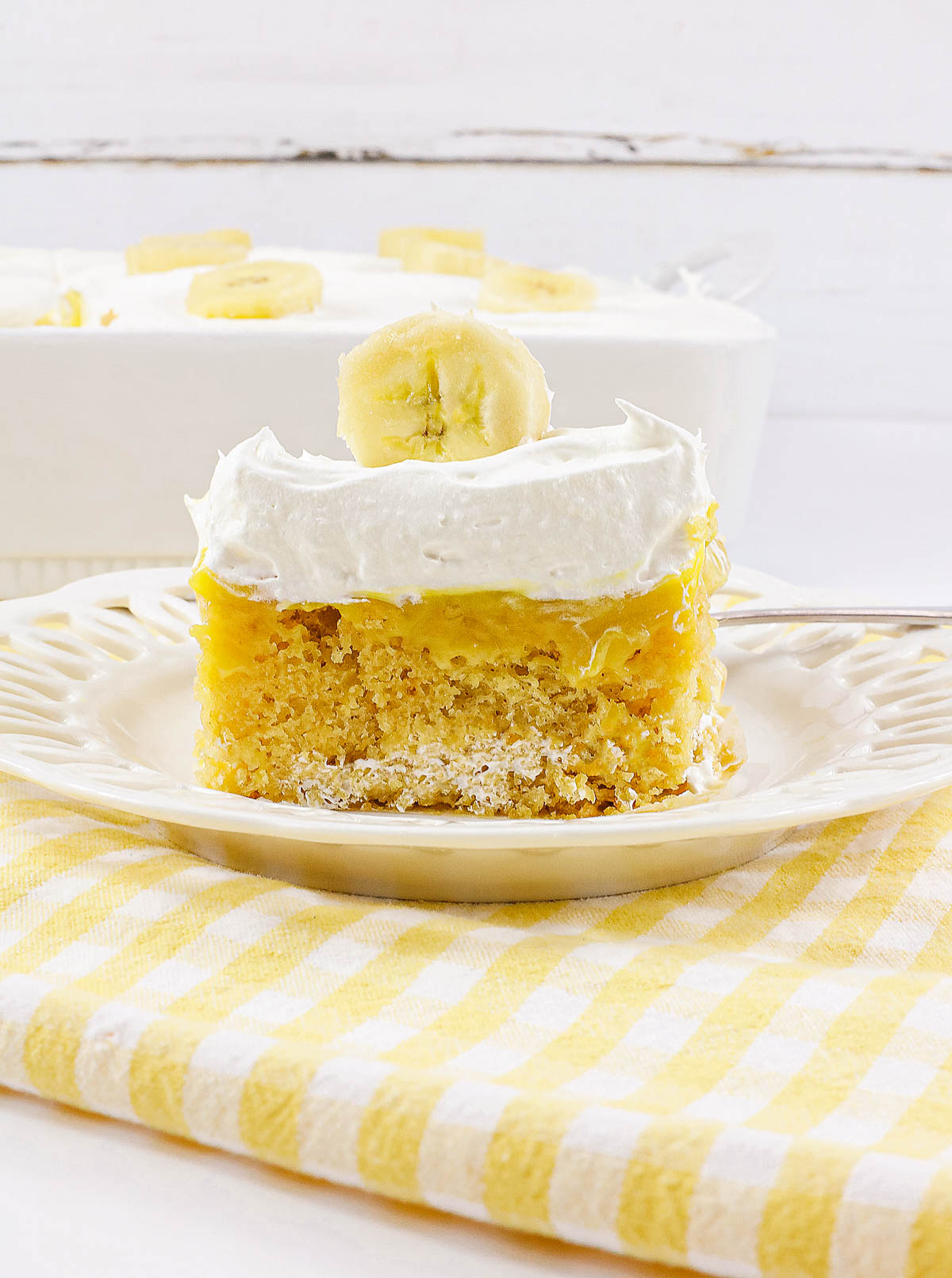 A slice of banana pudding poke cake with whipped topping and a banana slice on a white plate—made with creamy banana cream and no egg for a light, irresistible treat.