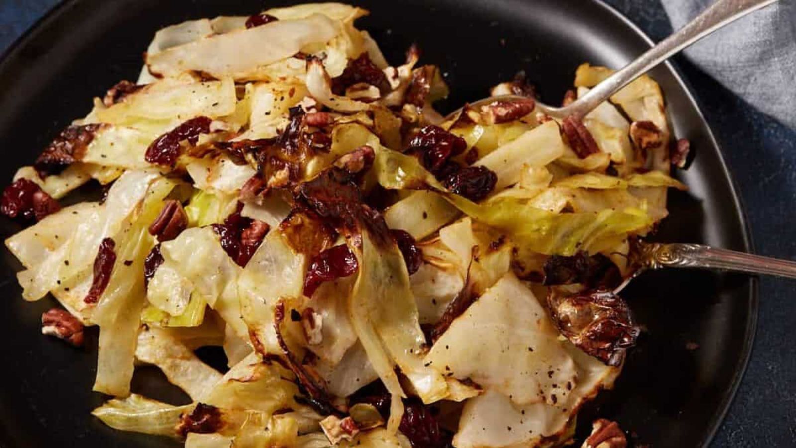 Roasted cabbage with dried cranberries and pecans served on a black plate with two forks.