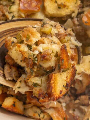 A wooden spoon scoops up baked stuffing with bread cubes and herbs from a casserole dish.