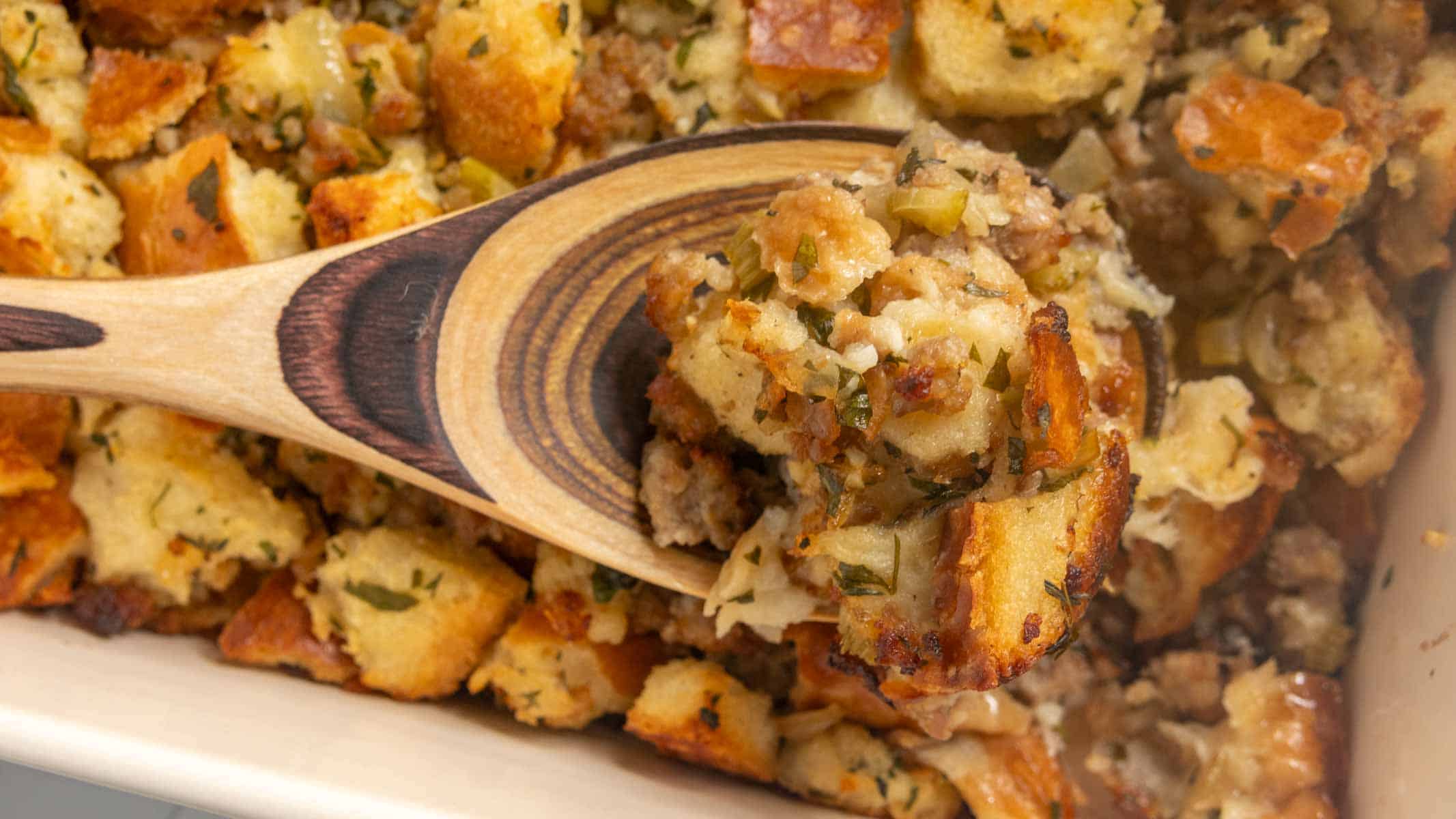 A wooden spoon scoops up baked stuffing with bread cubes and herbs from a casserole dish.