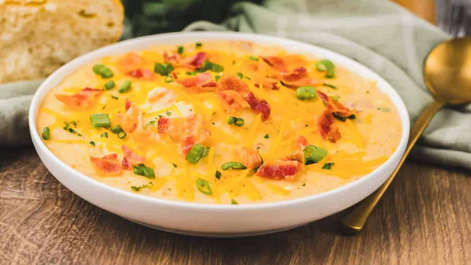 A bowl of creamy soup topped with shredded cheese, bacon bits, and chopped green onions on a wooden table.