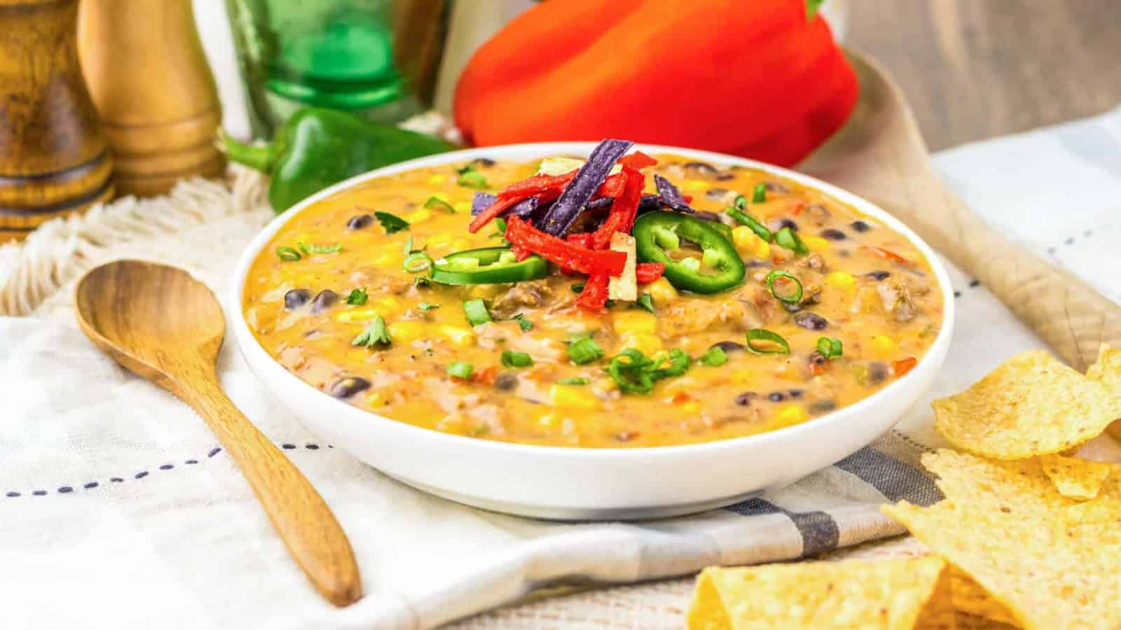 A bowl of cheesy soup with beans, peppers, jalapeños, and tortilla strips, next to tortilla chips.