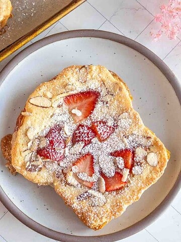 A plate with a slice of toast topped with strawberries and powdered sugar, next to a bowl of fresh strawberries.