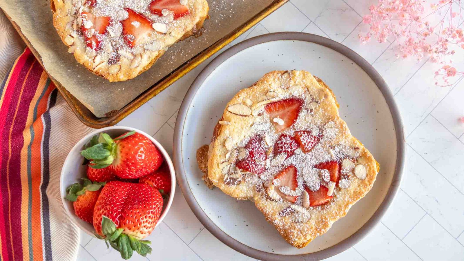 A plate with a slice of toast topped with strawberries and powdered sugar, next to a bowl of fresh strawberries.