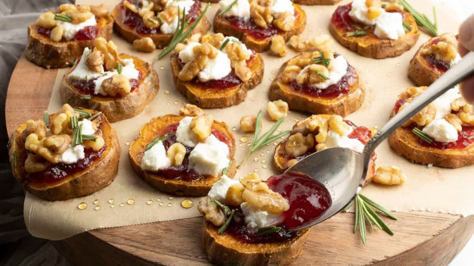 Slices of sweet potato topped with jam, cheese, walnuts, and rosemary are arranged on a wooden board.