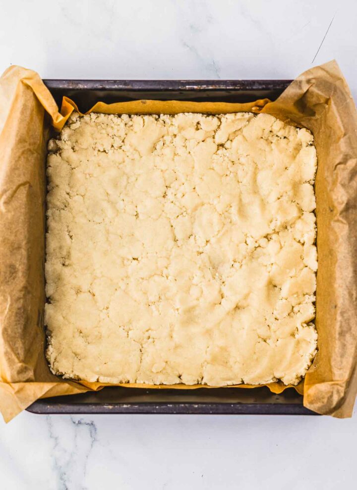 Unbaked crumbly dough for cherry pie crumb bars is pressed into a parchment-lined square baking pan on a marble surface, ready to start this delicious recipe.