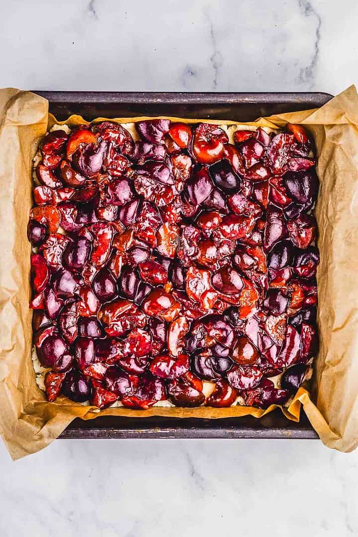 A square baking pan lined with parchment paper, filled with glossy roasted cherries on top of a crumb bar base&mdash;perfect for a homemade cherry pie recipe.