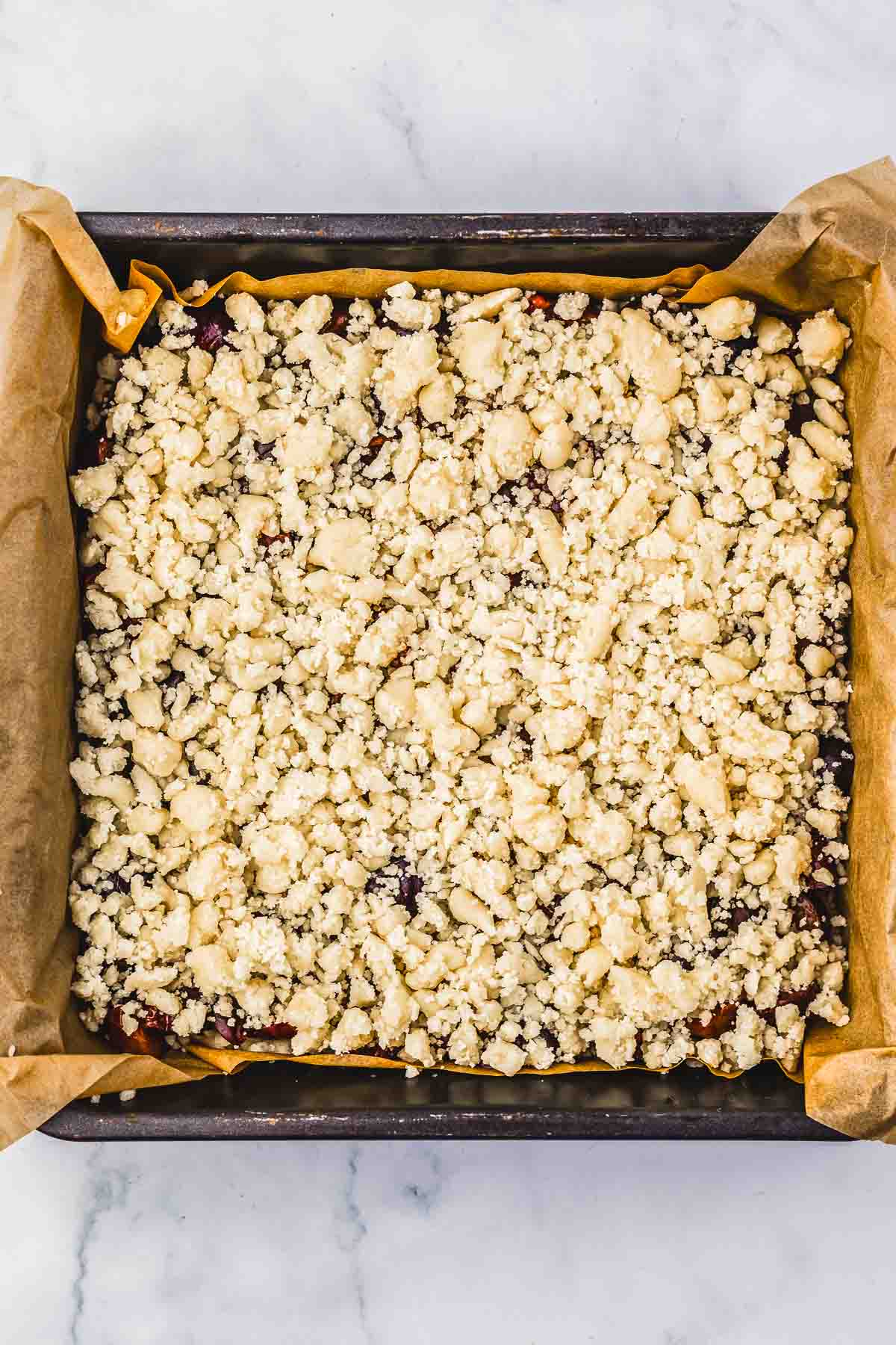 Unbaked cherry pie crumb bars in a parchment-lined square baking pan rest on a marble surface, ready for the next step in your favorite recipe.