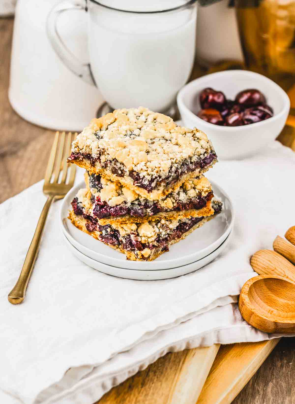 Three cherry crumble bars stacked on a plate with a fork, bowl of cherries, and milk in the background&mdash;these delicious crumb bars taste just like classic Cherry Pie in every bite.