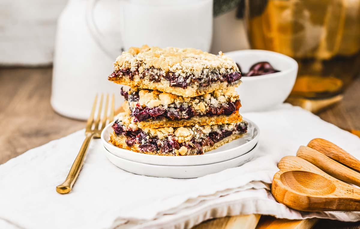 Three stacked crumb bars on a white plate, with a gold fork and wooden utensils beside them&mdash;this recipe is perfect for anyone craving a cherry pie-inspired treat.