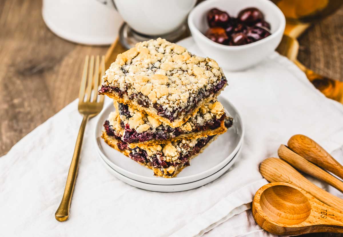 Three crumbly Cherry Pie Crumb Bars stacked on a white plate, with a fork and measuring spoons nearby on a napkin&mdash;perfect for anyone searching for their next favorite recipe.
