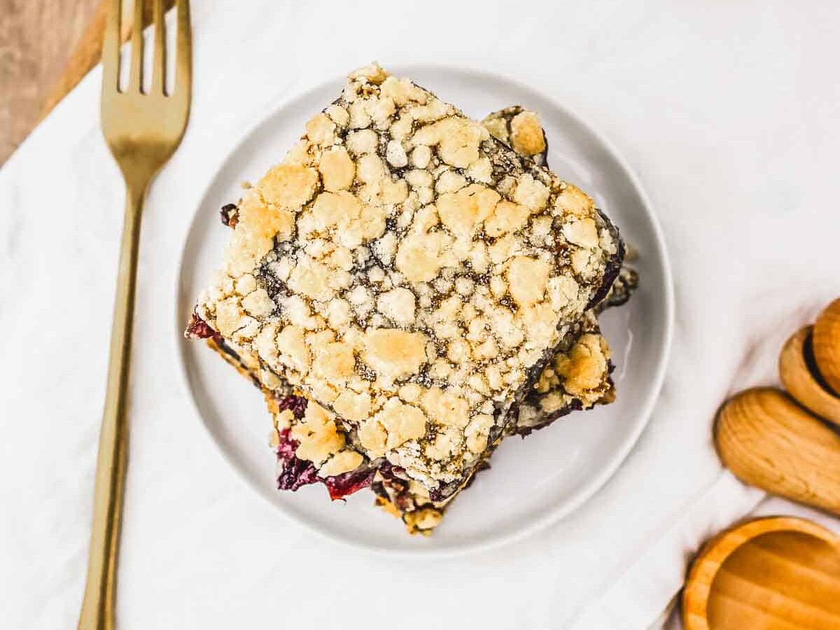 A crumbly square dessert bar&mdash;just like classic crumb bars&mdash;rests on a white plate beside a fork, with a bowl of dark cherries and a glass of milk, making it look like the perfect treat from a cherry pie recipe.