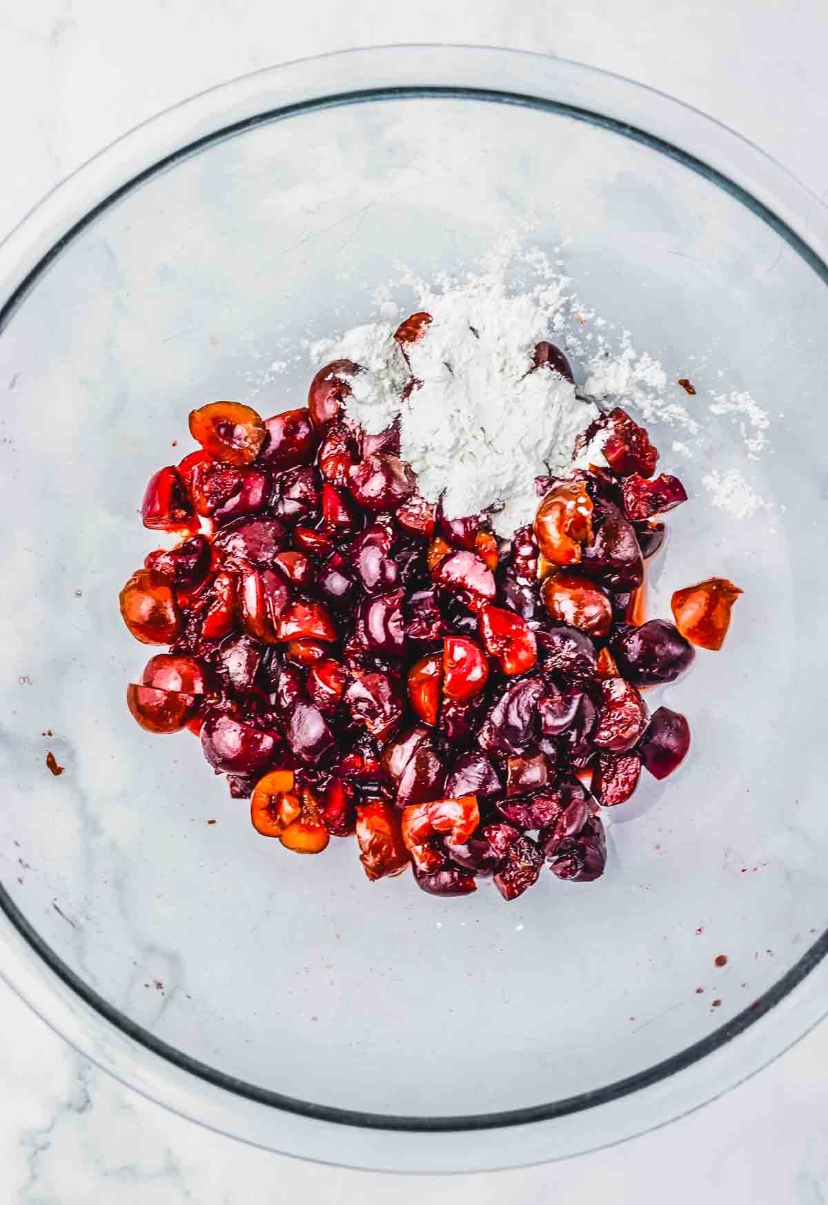 Halved cherries and a pile of white flour in a glass mixing bowl on a light surface, ready to be transformed into a delicious cherry pie or crumb bars recipe.