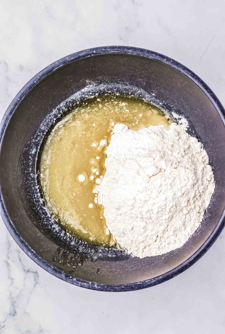 Black mixing bowl with melted butter and a pile of flour on a light marble countertop, ready for mixing up your favorite Crumb Bars recipe.
