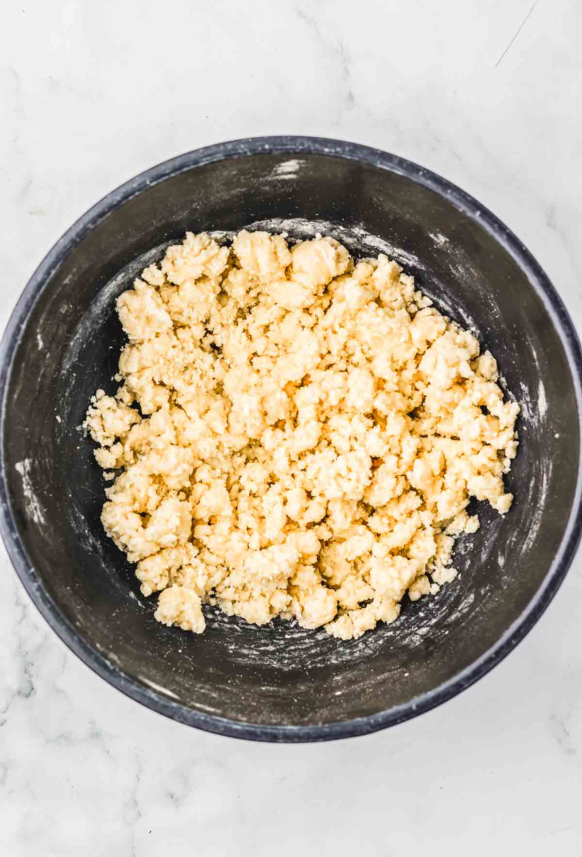 A black bowl filled with crumbly pie dough mixture, perfect for a Cherry Pie or Crumb Bars recipe, sits on a white marble surface.