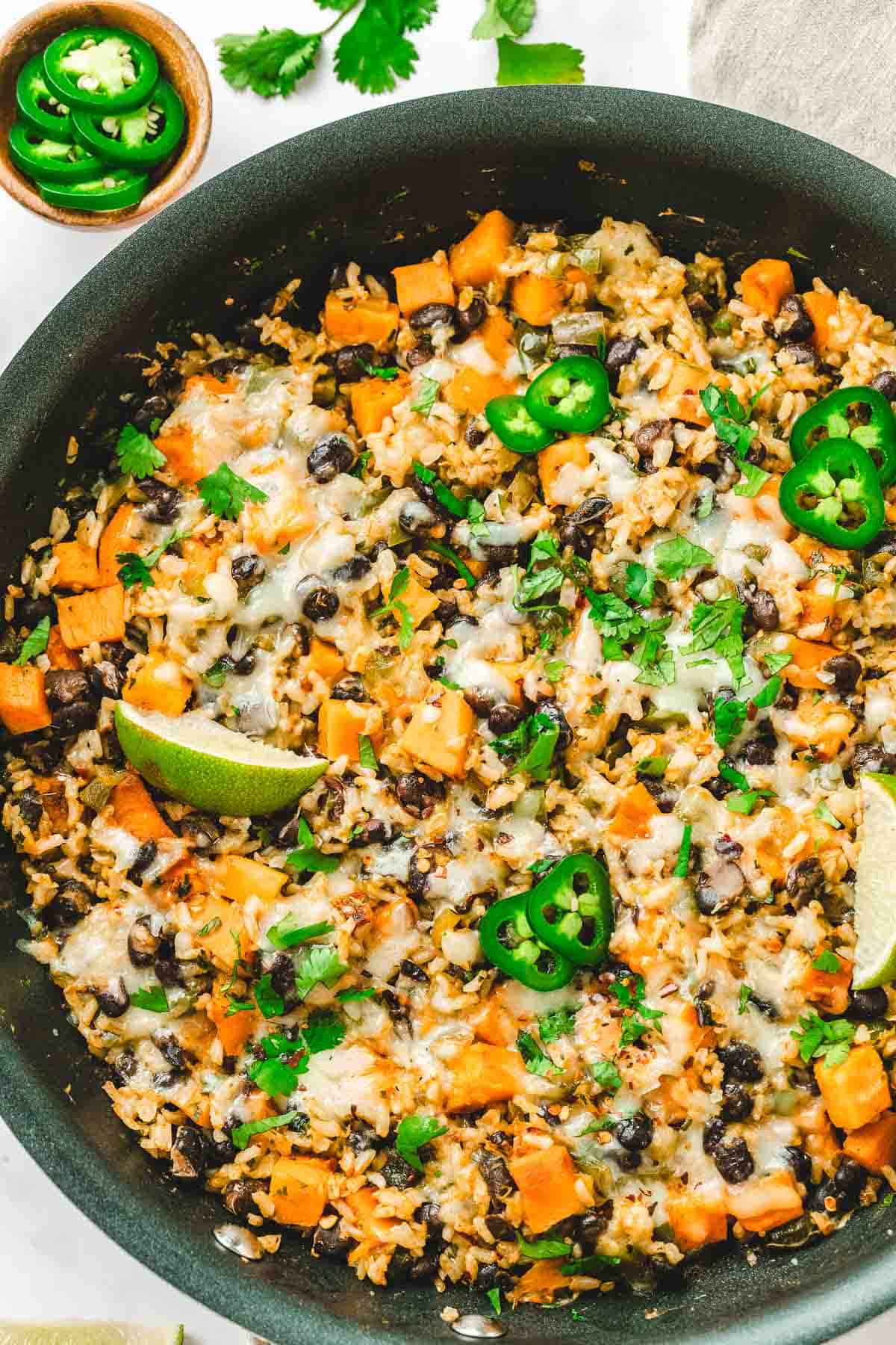A delicious Rice Skillet filled with cheesy rice, black beans, sweet potatoes, jalapeño slices, lime wedges, and fresh herbs.