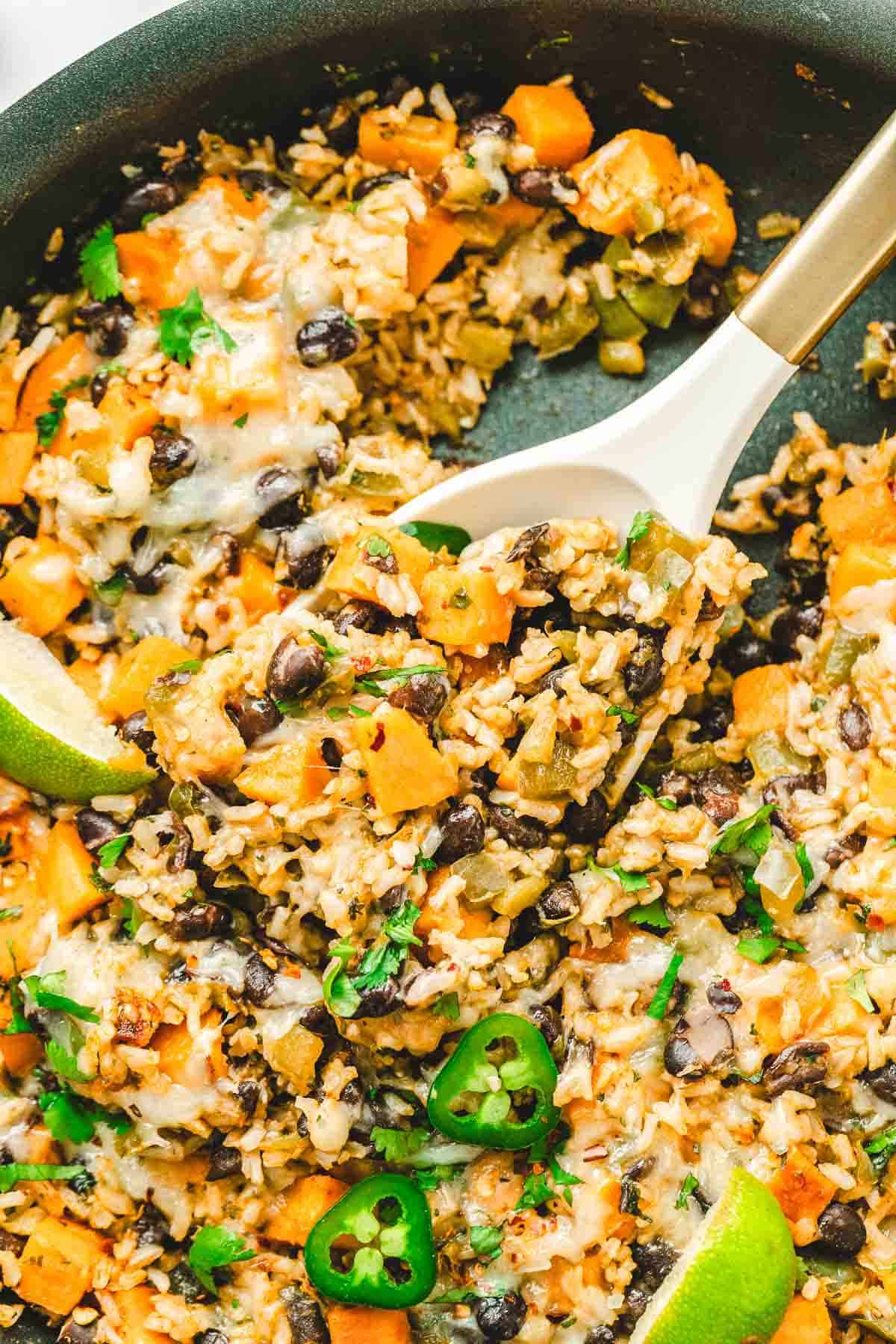 A spoon scooping cheesy rice skillet with black beans, Southwest sweet potato, jalapeños, and fresh herbs from a hot skillet.