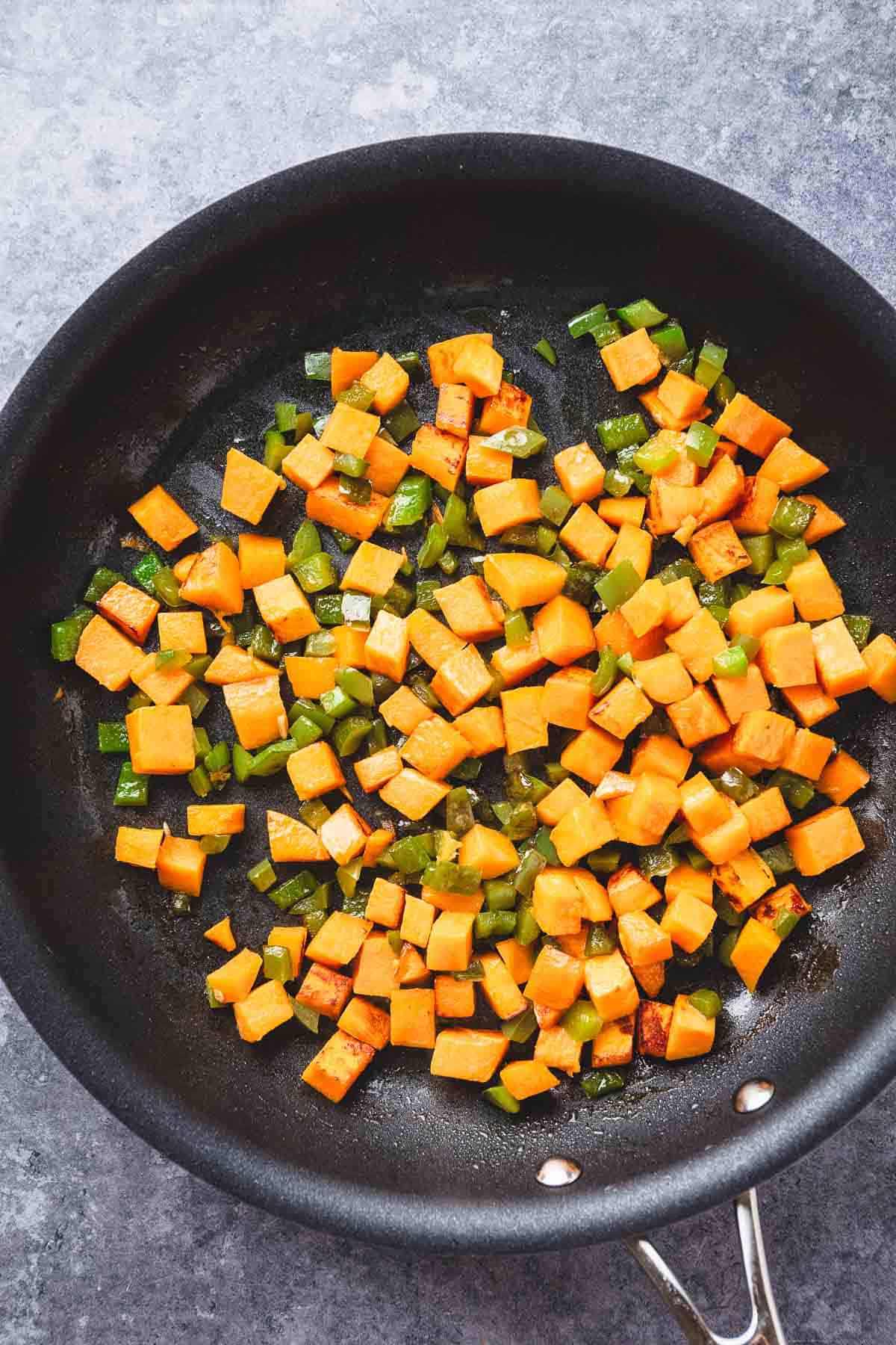 Diced sweet potatoes and green bell peppers sautéing in a black skillet for a tasty Southwest Sweet Potato and Black Bean dish on a gray countertop.