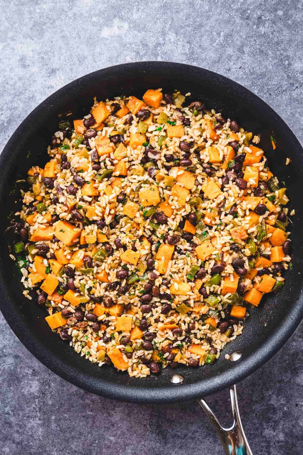A Southwest Sweet Potato and Black Bean Rice Skillet filled with rice, black beans, sweet potato, and chopped vegetables sits on a gray surface.