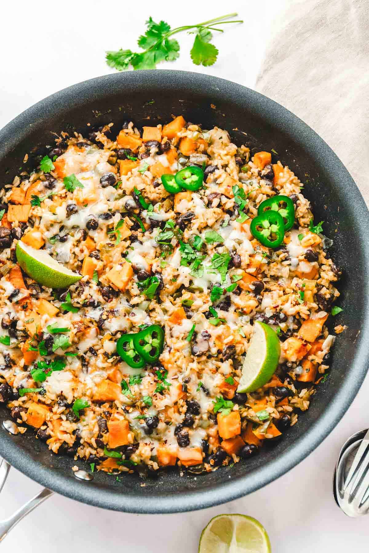 A Southwest Sweet Potato, Black Bean, and Rice Skillet featuring melted cheese, jalapeños, and lime wedges, all garnished with fresh cilantro.
