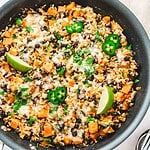 A skillet with rice, black beans, sweet potato, melted cheese, jalapeño slices, and lime wedges.