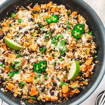 A skillet with rice, black beans, sweet potato, melted cheese, jalapeño slices, and lime wedges.