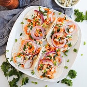 Four easy Chicken Tacos topped with shredded buffalo chicken, red onions, herbs, and cheese on a white plate, with extra toppings nearby—perfect for fans of simple Easy Recipes.
