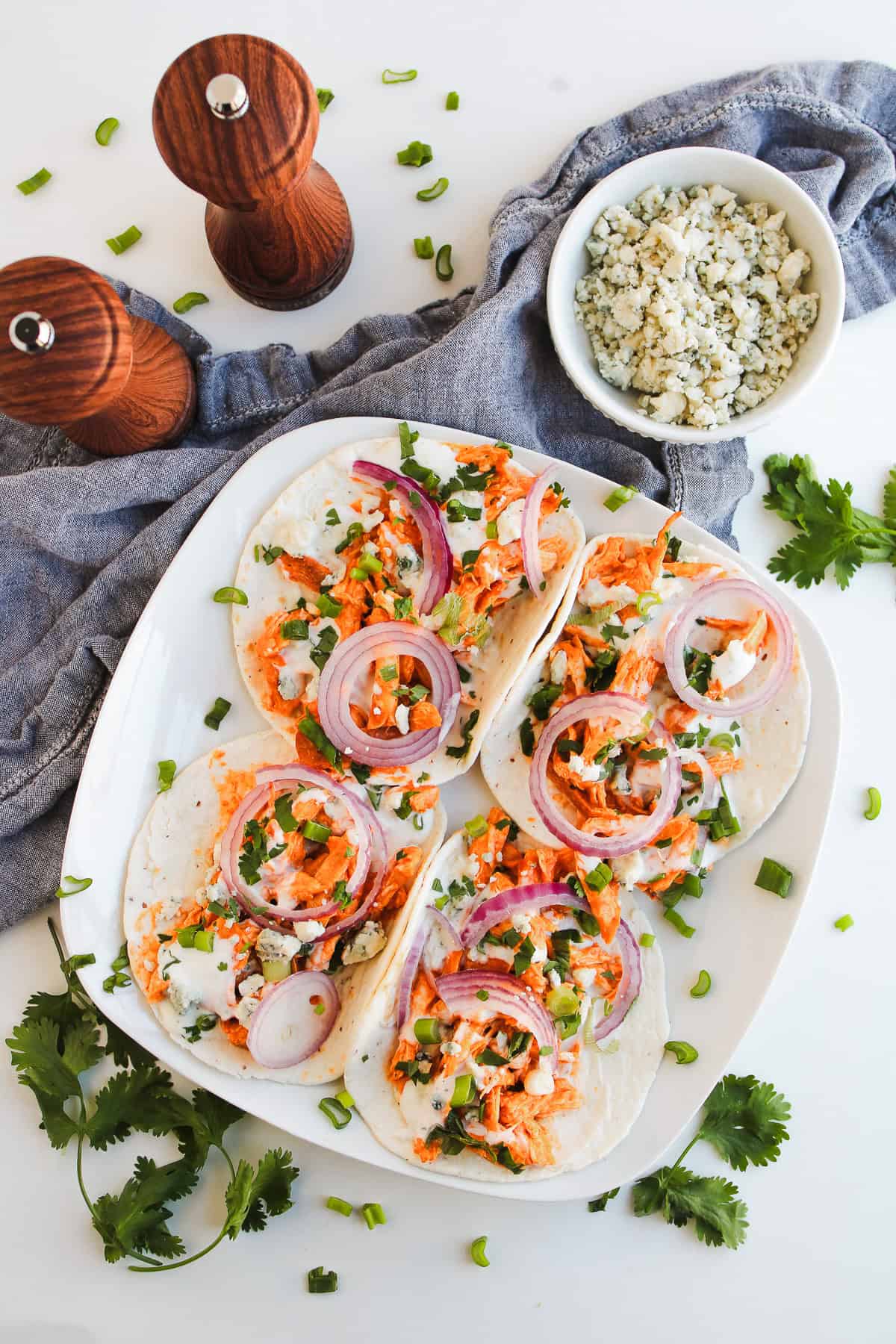 Four easy Chicken Tacos topped with shredded buffalo chicken, red onions, herbs, and cheese on a white plate, with extra toppings nearby—perfect for fans of simple Easy Recipes.