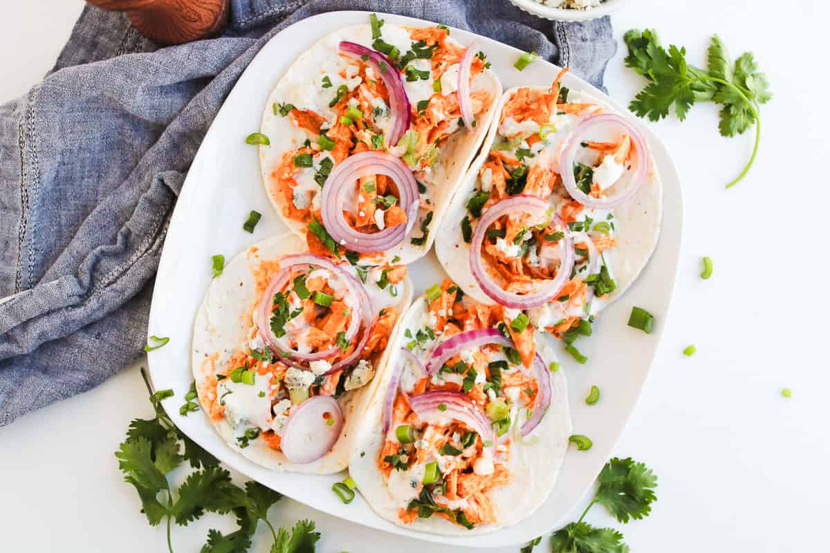 Four Buffalo Chicken Tacos with red onion, cilantro, and sauce on a white plate, surrounded by fresh cilantro.