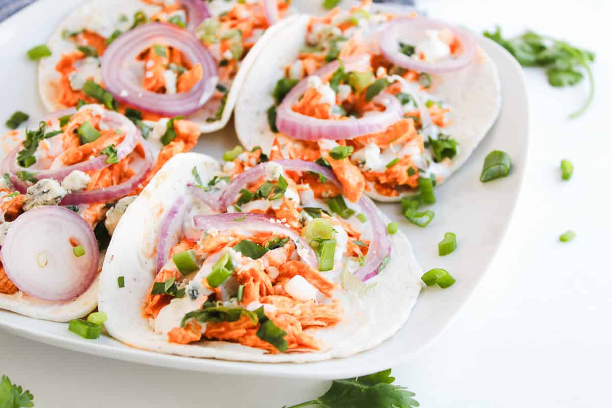 Soft tacos filled with Buffalo Chicken, sliced red onions, green onions, and a creamy sauce on a white plate—perfect for those who love easy recipes.