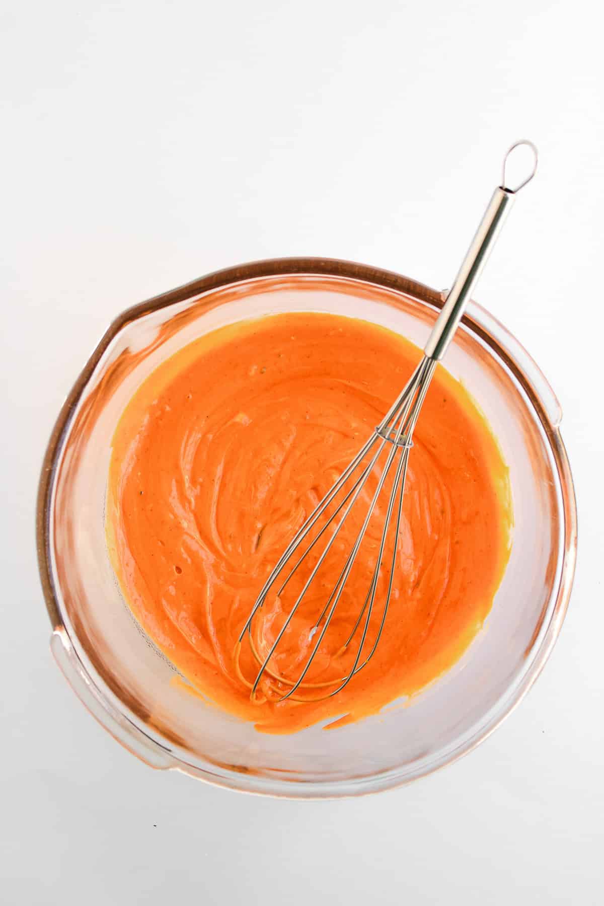 A glass bowl with orange Buffalo Chicken sauce and a metal whisk on a white background.