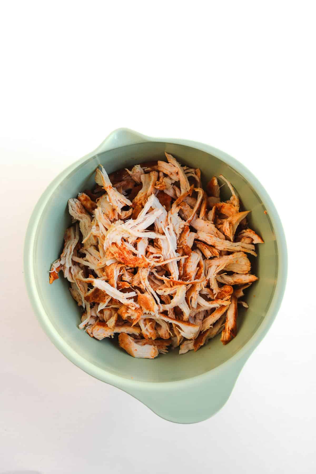 Shredded cooked chicken in a light green mixing bowl on a white background, perfect for making Buffalo Chicken Tacos or other easy chicken taco recipes.