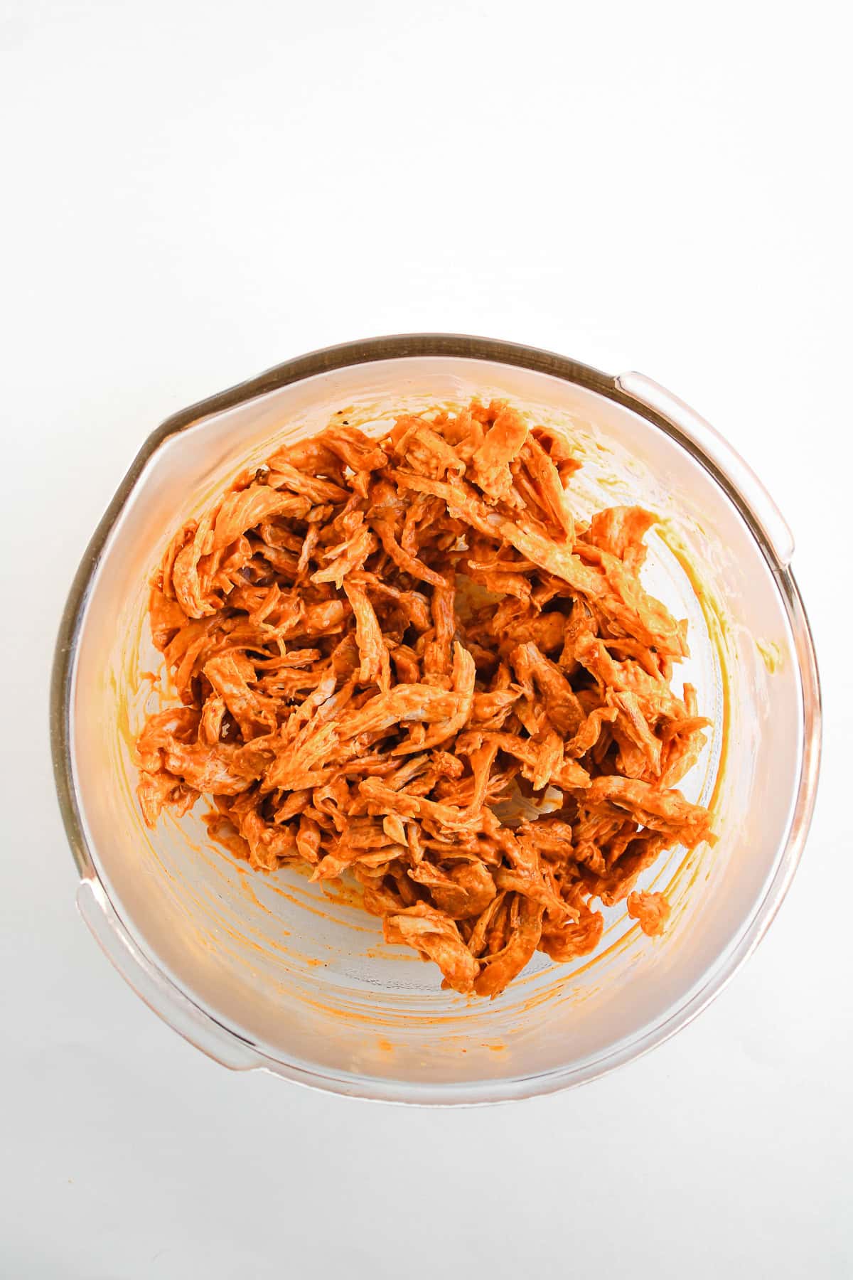 Shredded buffalo chicken in a glass bowl, coated with an orange-red sauce, perfect for easy recipes like chicken tacos, on a white background.