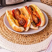 Two sausage and pepper sandwiches on a plate sit in front of a Crockpot and a potted plant—perfect for showcasing this easy crockpot sausage and peppers recipe.