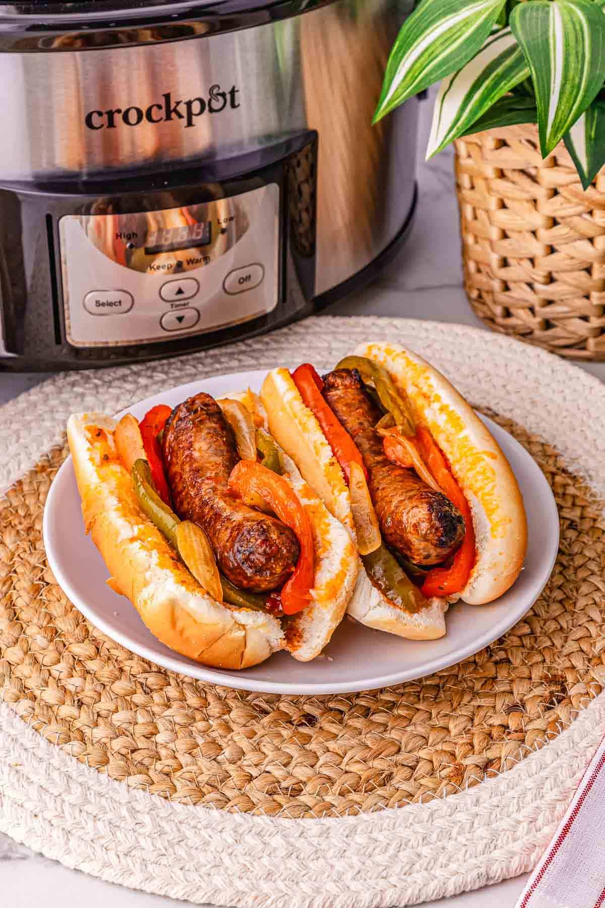 Two sausage and pepper sandwiches on a plate sit in front of a Crockpot and a potted plant&mdash;perfect for showcasing this easy crockpot sausage and peppers recipe.