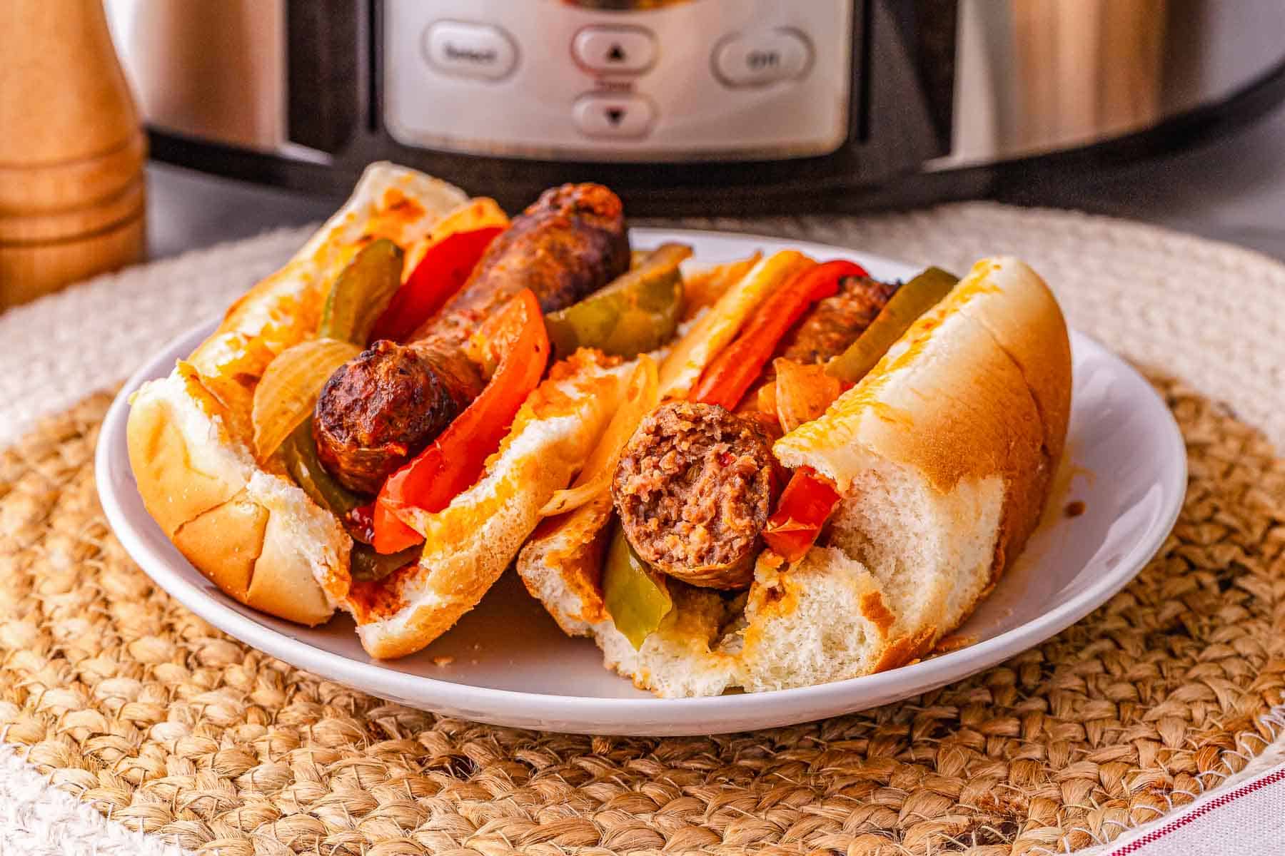 Two sausage and pepper sandwiches with onions on a white plate in front of a slow cooker, perfect for enjoying this classic crockpot sausage and peppers recipe.