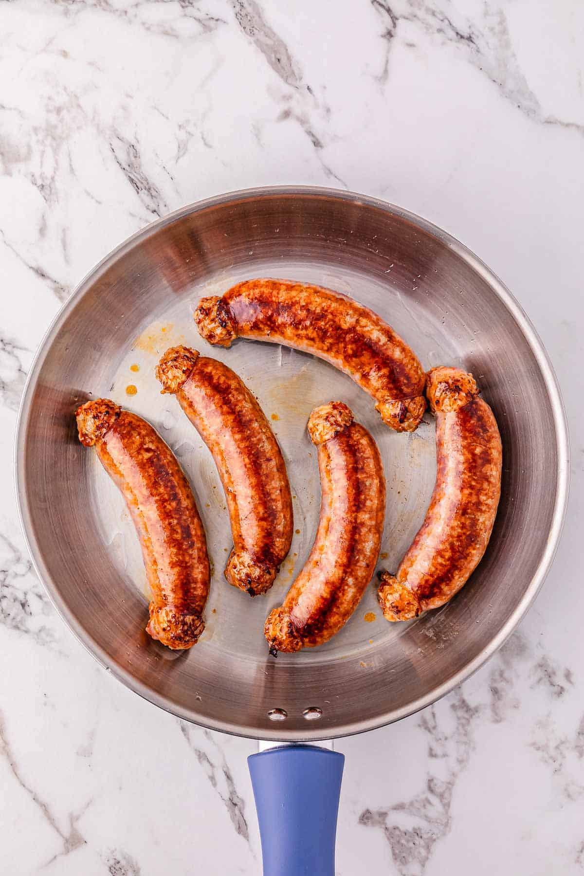 Five browned sausages cooking in a stainless steel skillet on a marble countertop.