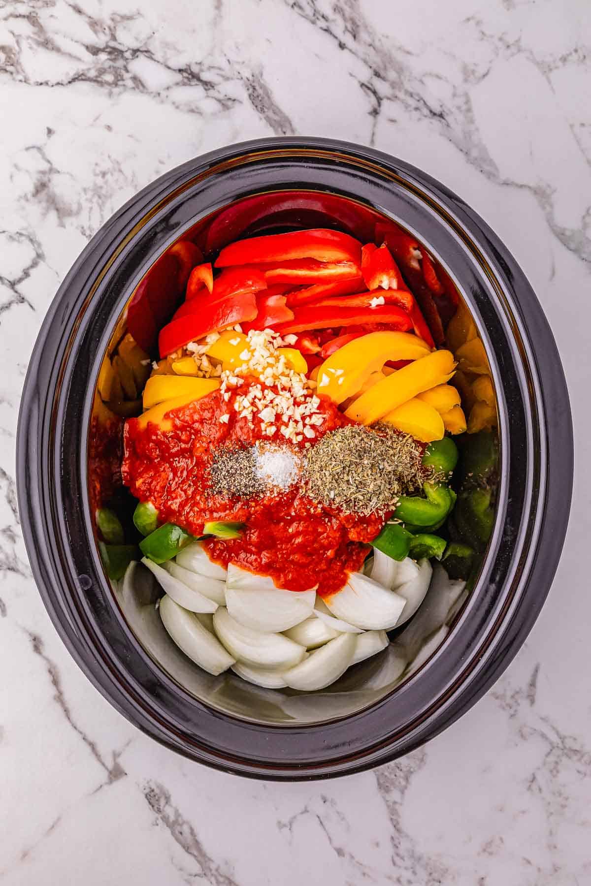 A crockpot sausage and peppers recipe featuring sliced onions, bell peppers, tomato sauce, minced garlic, and spices in a slow cooker on a marble surface.
