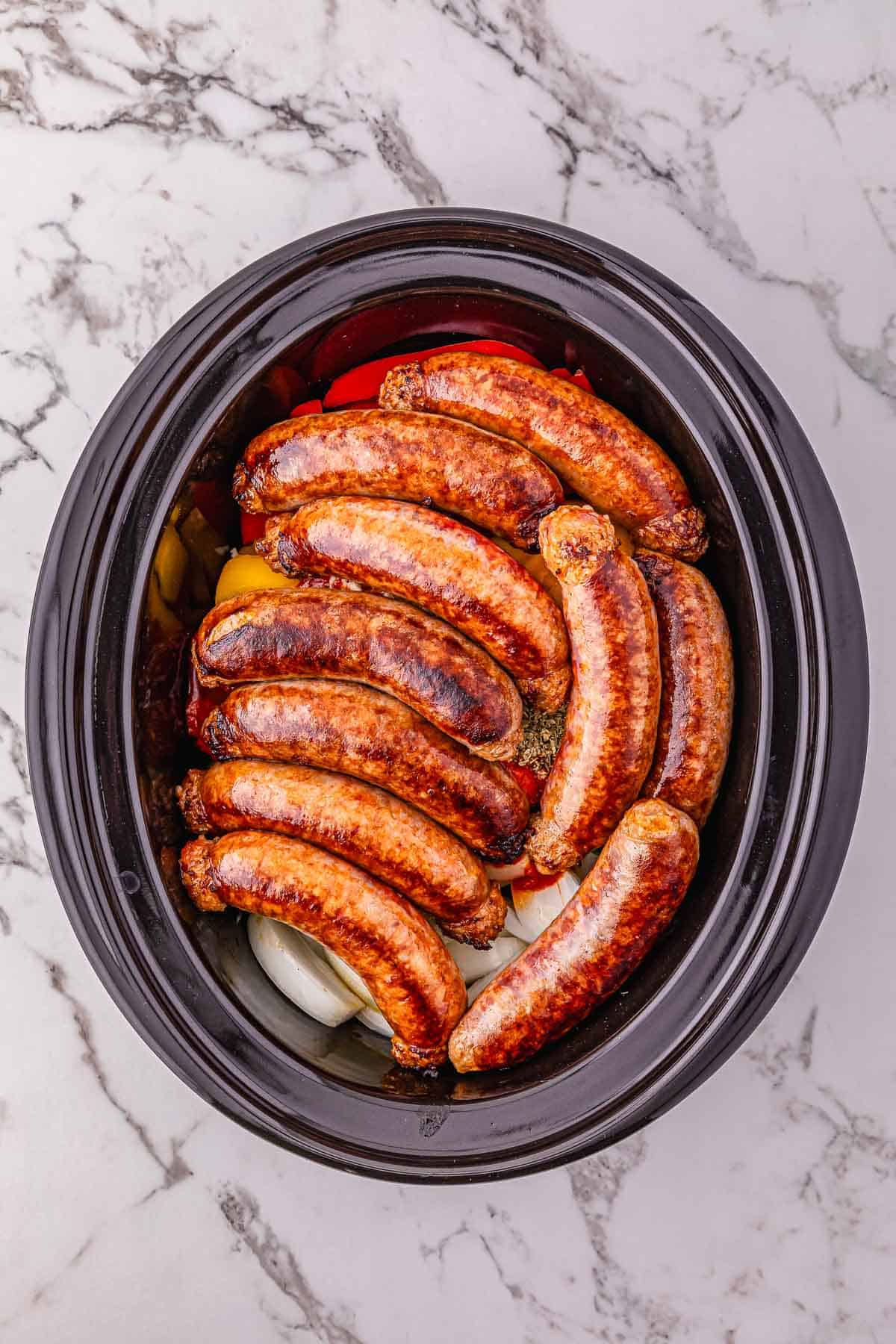 Sausages arranged in a slow cooker on top of sliced onions and peppers on a marble countertop&mdash;an easy crockpot sausage and peppers recipe perfect for effortless weeknight dinners.
