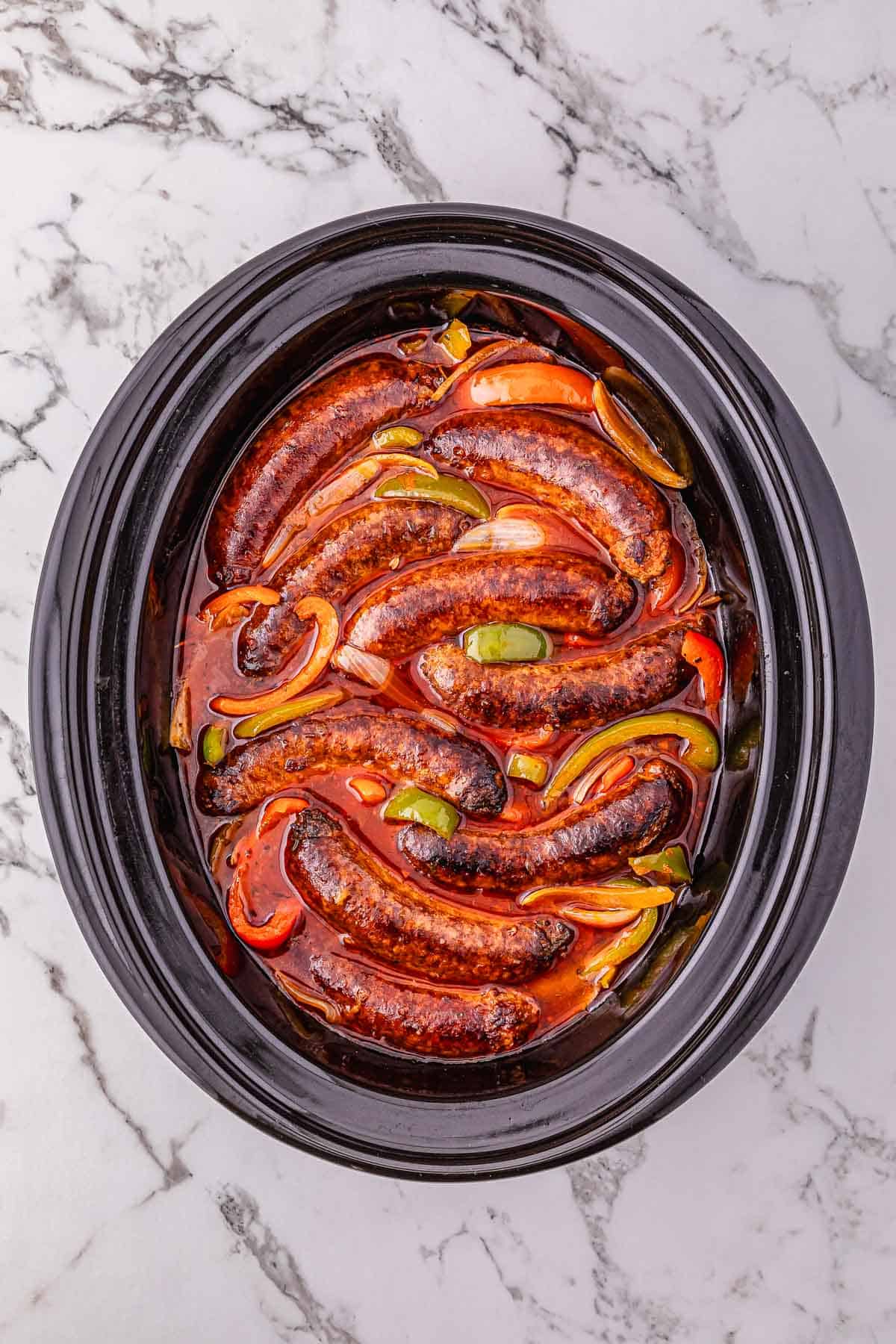 Crockpot sausage and peppers with onions in a rich red sauce fill the slow cooker, ready to serve on a marble surface.