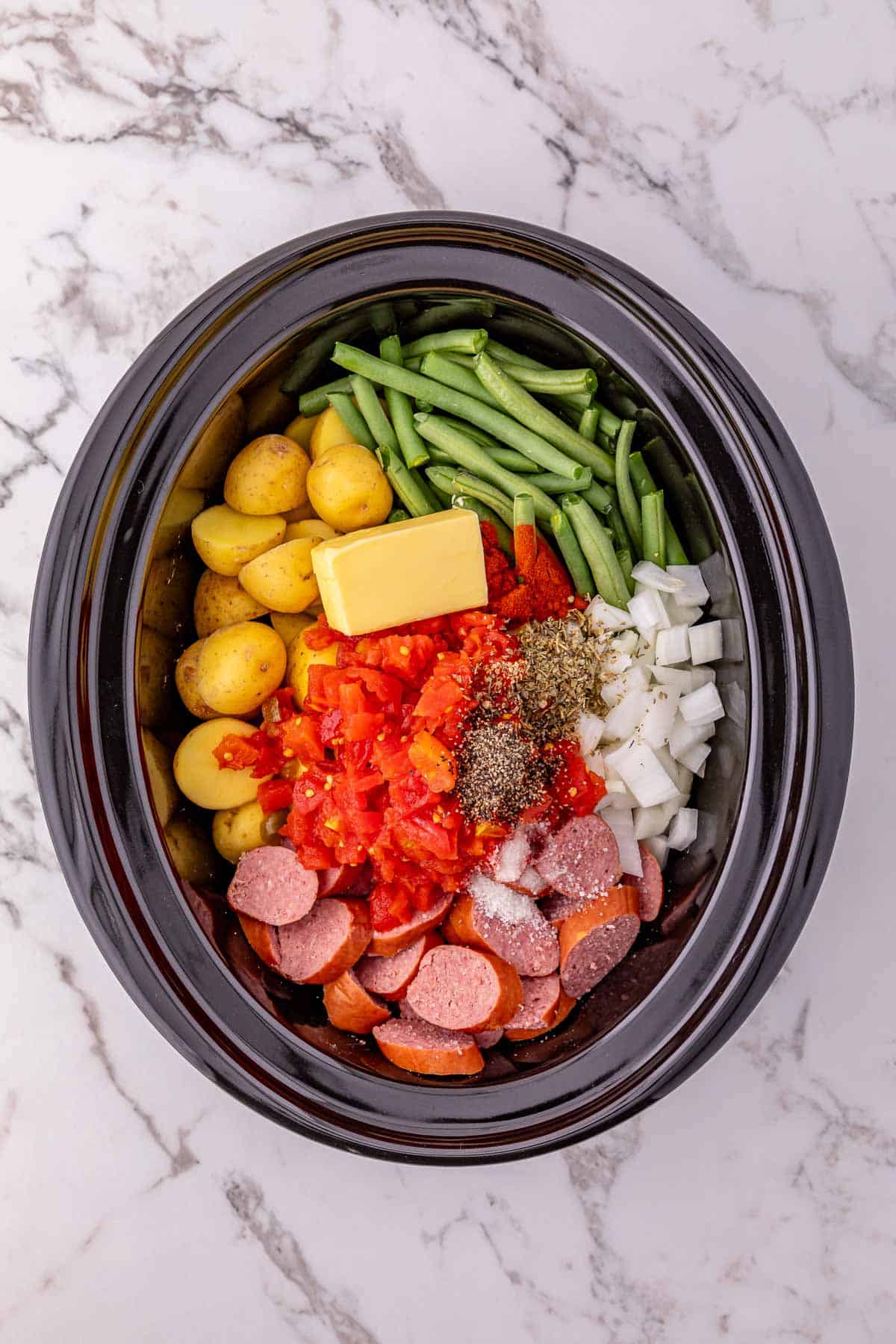 Crockpot sausage and potatoes simmer with green beans, onions, diced tomatoes, spices, and butter in a slow cooker on a marble surface.
